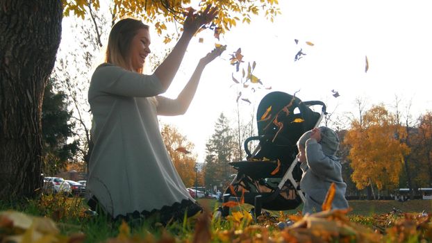 Young mother with her laughing little baby throwing leaves in the air in autumn park. Mid shot
