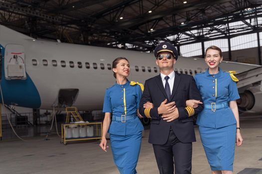 Male pilot in uniform and aviator sunglasses walking together with two air stewardesses near airplane in hangar