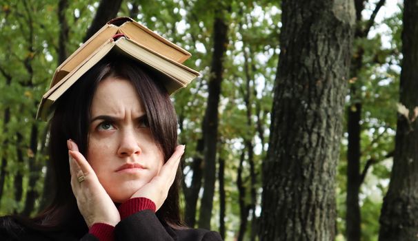 Portrait of a young and funny brunette in the park holding an open book on her head. Learning is fun. Woman balancing with books on her head. The student is tired of reading