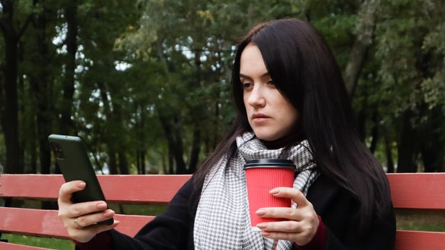 Beautiful young caucasian woman in a coat with a paper cup of takeaway coffee taking a selfie or taking pictures of herself for a blog with a smartphone outdoors in a park sitting on a bench