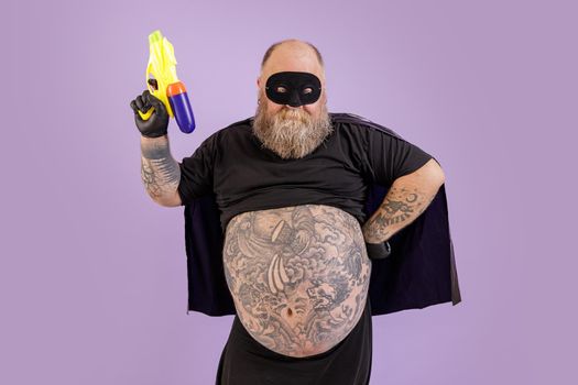 Mature bearded man with large tattooed abdomen wearing carnival costume with cape and mask holds toy blaster posing on purple background in studio