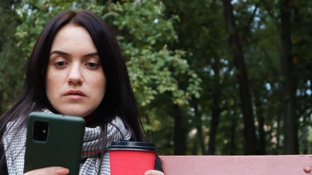 Beautiful young caucasian woman in a coat with a paper cup of takeaway coffee taking a selfie or taking pictures of herself for a blog with a smartphone outdoors in a park sitting on a bench