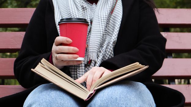 Young woman in jeans, coat and scarf, on a park bench. A woman is reading a book and drinking coffee or other hot drink outdoors alone. Close-up. The concept of honor, study, leisure and recreation