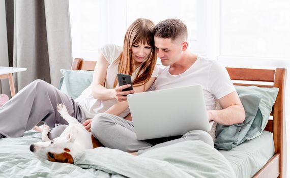Young couple in the bed with gadgets and cute dog. Beautiful woman showing her husband holding laptop something on smartphone. Family morning with pet and technologies