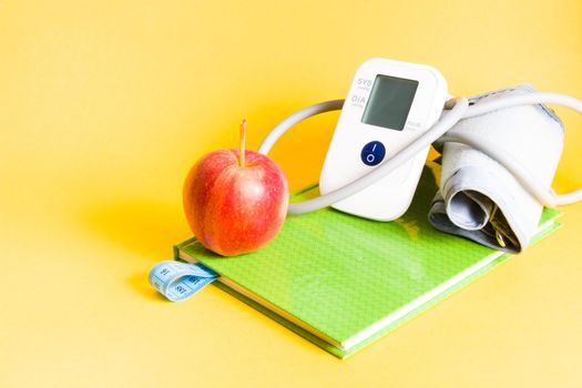 blood pressure monitor on a green notebook, a red apple and a notebook with a bookmark from a measuring blue tape, weight loss and health care, proper nutrition concept