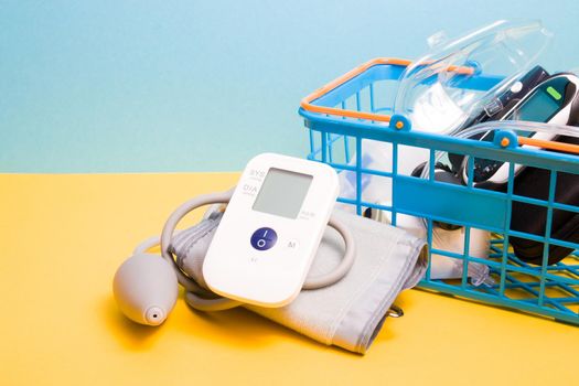 measure blood pressure monitor lies next to a small blue shopping basket in which there is a glucose meter and a nebulizer for inhalation yellow-blue background copy space, pharmacy concept