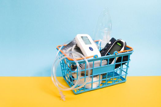 medical devices for home use in a small blue shopping basket, yellow-blue background copy space, glucose meter, inhalation nebulizer, measure blood pressure monitor