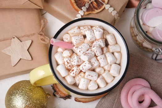 yellow cup with cocao decorated with marshmallows on a stand made of saw cut wood, a jar of marshmallows, a spruce branch and Christmas gifts on the background