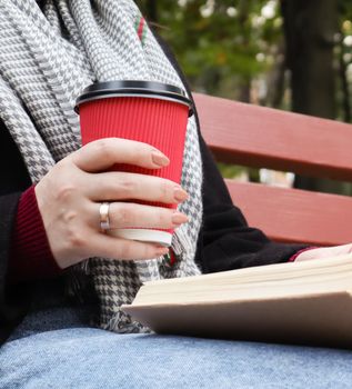 Young woman in jeans, coat and scarf, on a park bench. A woman is reading a book and drinking coffee or other hot drink outdoors alone. Close-up. The concept of honor, study, leisure and recreation