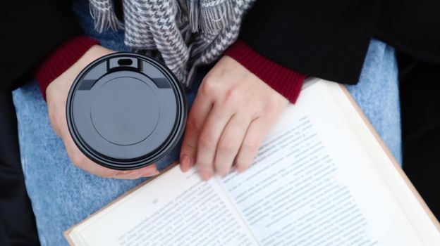 Young woman in jeans, coat and scarf, on a park bench. A woman is reading a book and drinking coffee or other hot drink outdoors alone. Close-up. View from above. The concept of honor, leisure