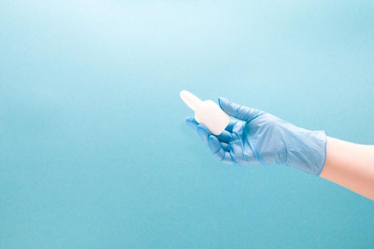 a female hand in a blue disposable medical glove holds a white plastic bottle with drops for the nose, blue background copy space, nasal drops, medicine for the nose