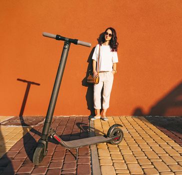 Smiling beautiful young woman in white clothes standing near the wall in city street, electric scooter in foreground.