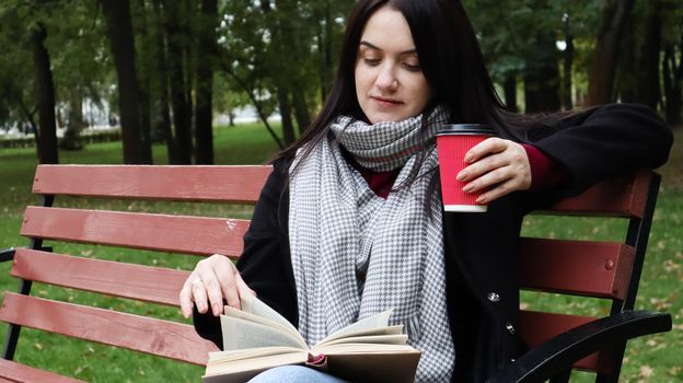 Young woman in jeans, coat and scarf, on a park bench. A woman is reading a book and drinking coffee or other hot drink outdoors alone