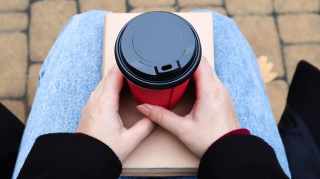Young woman in jeans, coat and scarf, on a park bench. A woman is reading a book and drinking coffee or other hot drink outdoors alone. Close-up. View from above. The concept of honor, leisure