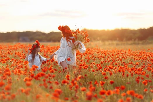 Little girl with redhead mother in white dresses and wreathes walking with bouquet of poppies on poppy field at warm summer sunset