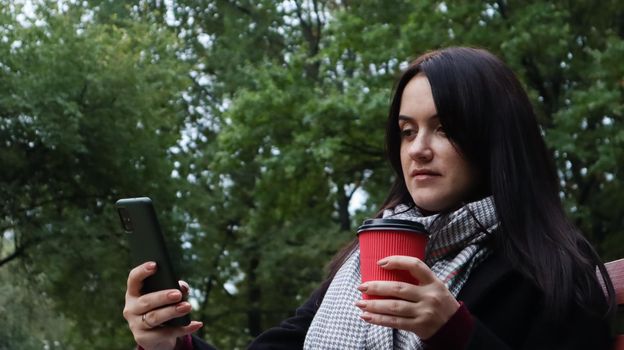 Beautiful young caucasian woman in a coat with a paper cup of takeaway coffee taking a selfie or taking pictures of herself for a blog with a smartphone outdoors in a park sitting on a bench