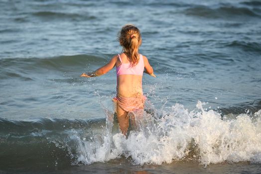 Girl in pink swimsuit standing on the beach and looking at the morning sea with waves. Child on the sea beach and beautiful skyline.