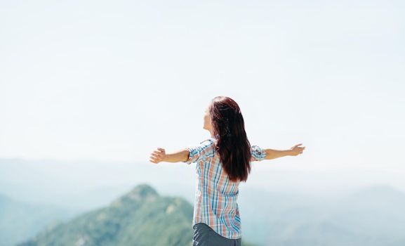 Happy young woman standing with raised arms high in mountains and enjoying view of nature in summer outdoor.