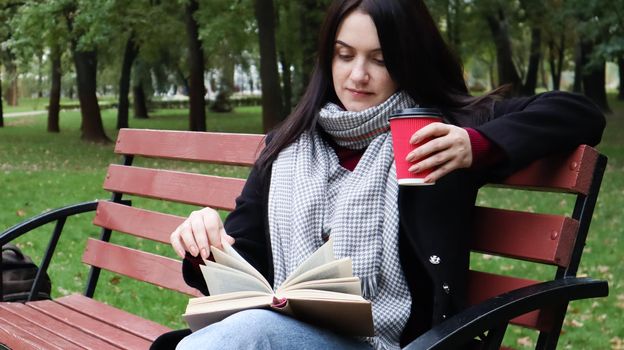 Young woman in jeans, coat and scarf, on a park bench. A woman is reading a book and drinking coffee or other hot drink outdoors alone