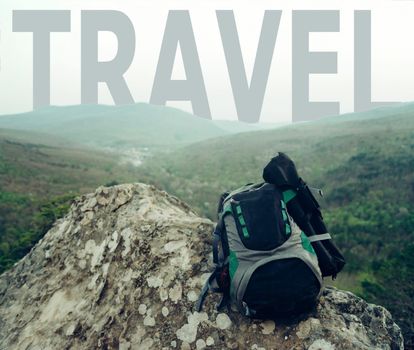 Backpack on peak of cliff in front of word travel on sky over mountain ridge.