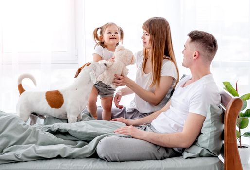 Sunny family mornents. Beautiful parents with their daughter and cute dog staying in the bed and enjoying time together. Mother, father and child smiling and doggy pet standing close to them