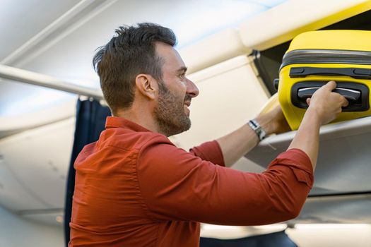 Side view of smiling male passenger holding suitcase and putting it on shelf in an airplane. Trip concept