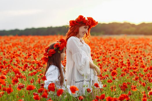 Little girl with redhead mother in white dresses and wreathes poses with bouquet of poppies on poppy field at warm summer sunset