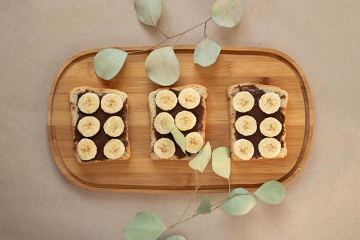 Three banana white bread toasts smeared with chocolate butter that lie on a cutting board with a sprig of leaves on craft paper background. top view with area for text