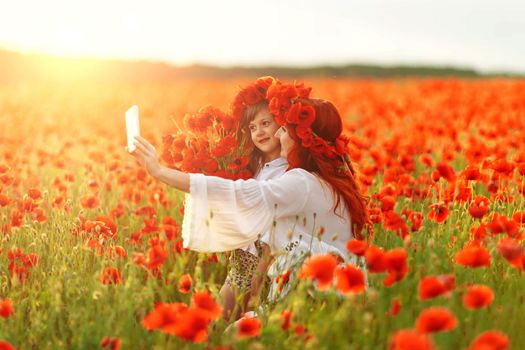 Little girl with redhead mother in white dresses and wreathes make selfie with bouquet of poppies on poppy field at warm summer sunset