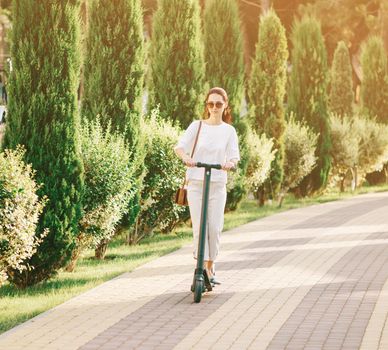 Stylish beautiful young woman riding to work an electric scooter in summer park. Concept of ecological transport.