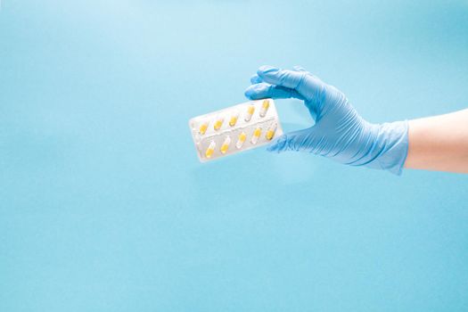 female hand in a blue disposable medical glove holds a blister with yellow capsule capsulim on a blue background copy space
