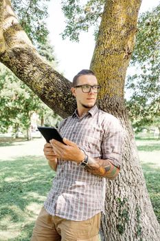 Handsome casual style young man standing with digital tablet near the tree in park.