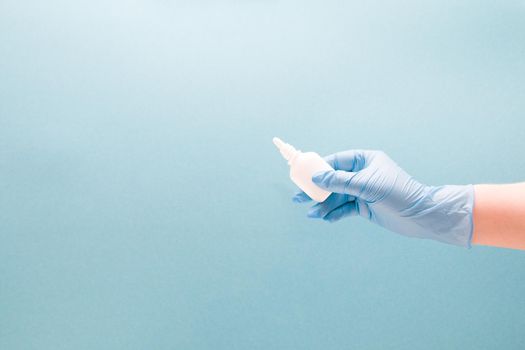 a female hand in a blue disposable medical glove holds a white plastic bottle with drops for the nose without a cap, blue background copy space, nasal drops, medicine for the nose