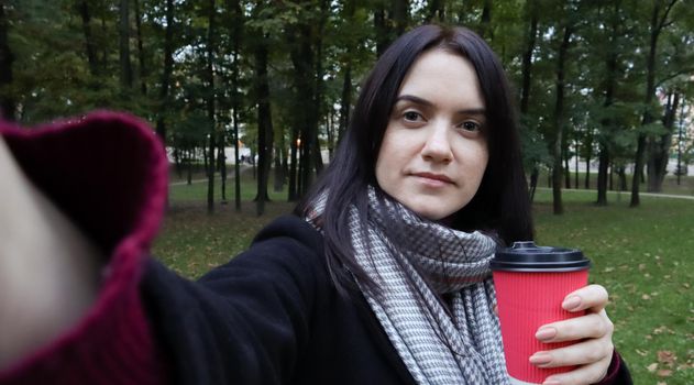 Beautiful young caucasian woman in a coat with a paper cup of takeaway coffee taking a selfie or taking pictures of herself for a blog with a smartphone outdoors in an autumn park
