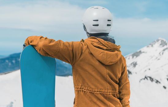 Young woman wearing in helmet standing with snowboard and enjoying view of mountains in winter, rear view