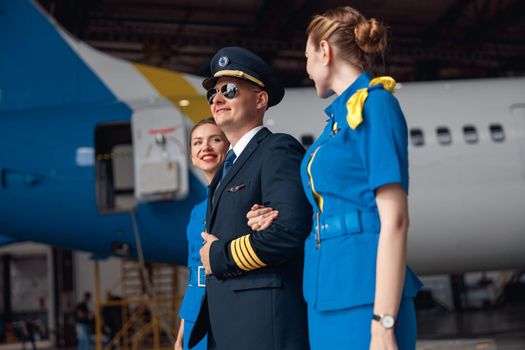 Confident pilot in uniform and aviator sunglasses walking together with two air stewardesses in blue uniform in front of big passenger airplane in airport hangar. Aircraft, occupation concept