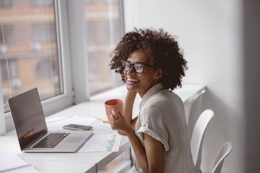 Smiling multiethnic woman wearing glasses and sitting at the desk with laptop while enjoying coffee