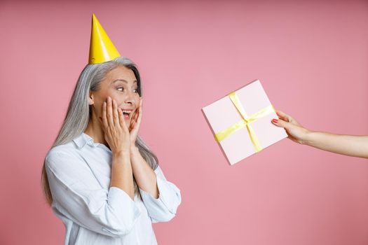 Person gives present to surprised middle aged Asian woman with silver hair and yellow party on pink background in studio