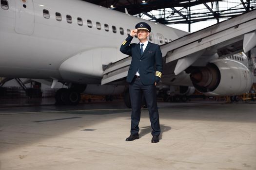 Full length shot of confident pilot in uniform looking away, adjusting his hat, standing in front of big passenger airplane in airport hangar. Aircraft, occupation, transportation concept