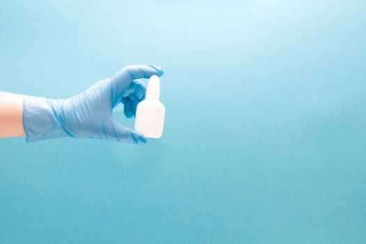 a female hand in a blue disposable medical glove holds a white plastic bottle with drops for the nose, blue background copy space, nasal drops, medicine for the nose