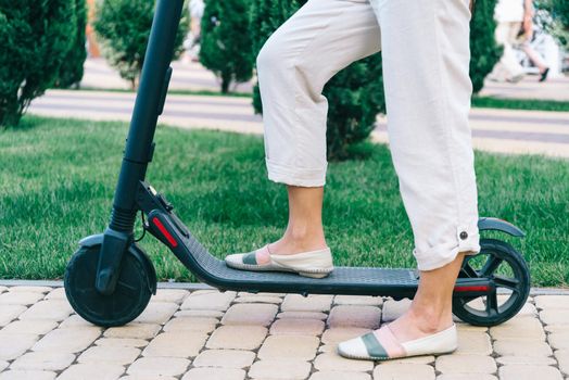 Unrecognizable young woman riding an electric scooter in city park in summer, view of legs.