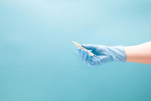 female hand in a blue disposable medical glove holds a mercury thermometer, blue background copy space
