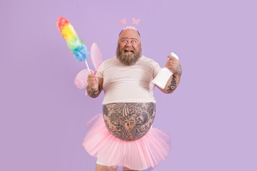 Happy middle aged bearded man with overweight in funny fairy costume holds pp duster and detergent bottle on purple background in studio