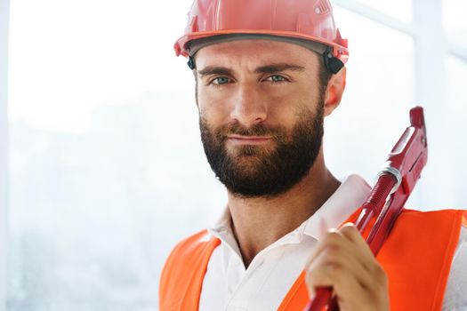 Young male plumber in workwear holding pipe wrench on a construction site indoors