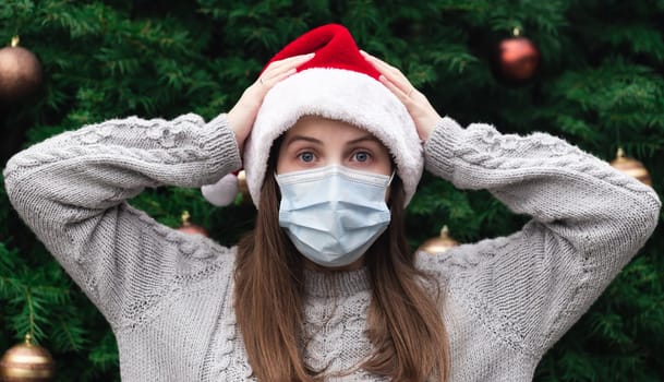 Shocked or surprised Christmas. Close up Portrait of woman wearing a santa claus hat and medical mask with emotion. Against the background of a Christmas tree. Coronavirus pandemic