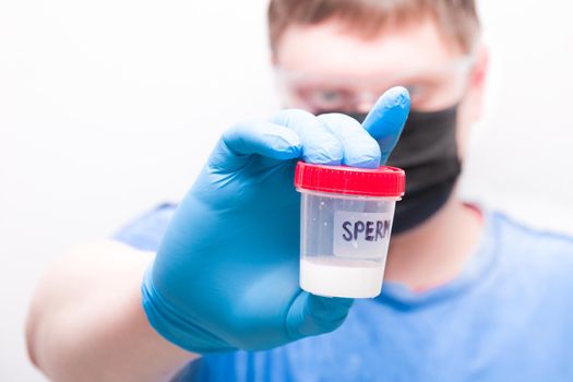 a man in goggles and a medical black mask holds a jar of sprerm in his hand. a hand in a blue disposable glove, a test jar with the inscription "sperm", blurred background, copy space