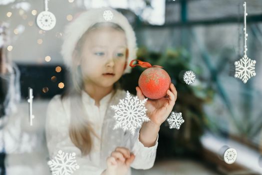 A girl wearing a santa claus hat looks at snowflakes on a window