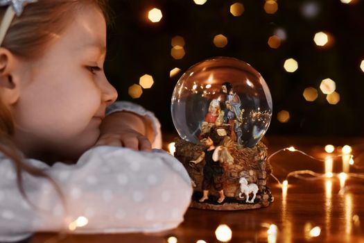 Girl looking at a glass ball with a scene of the birth of Jesus Christ near christmas tree