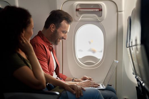 Side view of cheerful caucasian man smiling while using laptop computer, sitting on the airplane. Transportation concept