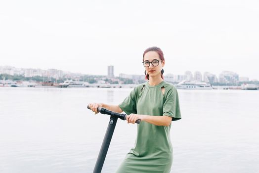 Beautiful young woman standing with electric scooter on background of sea bay and city, looking at camera.